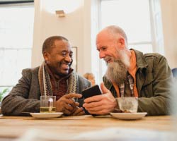 Two men in a coffee shop look at a phone and smile at each other.