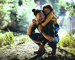 A man crouched on a hiking trail with a smiling child on his back.