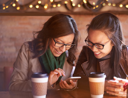 A pair of women in a coffee shop laugh while looking at a phone.