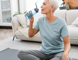 A woman sits on a yoga mat in her living room, drinking from a water bottle.